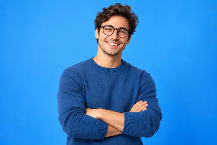 Freundlicher junger Berufstätiger mit Brille und blauem Pullover, der mit verschränkten Armen vor einem leuchtend blauen Studiohintergrund lächelt.